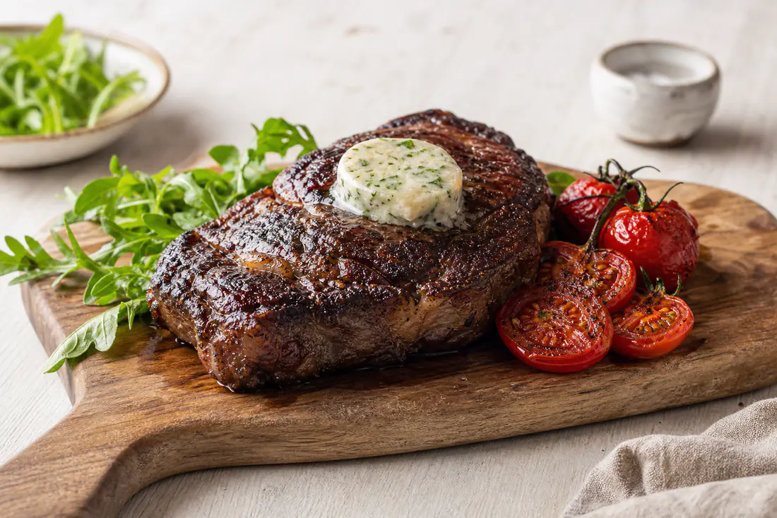 A thick ribeye steak with a dark crust and garlic butter on a board with roasted tomatoes and rocket