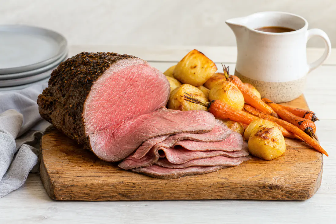 Thin slices of pink roast beef on a board with roast potatoes, carrots and gravy