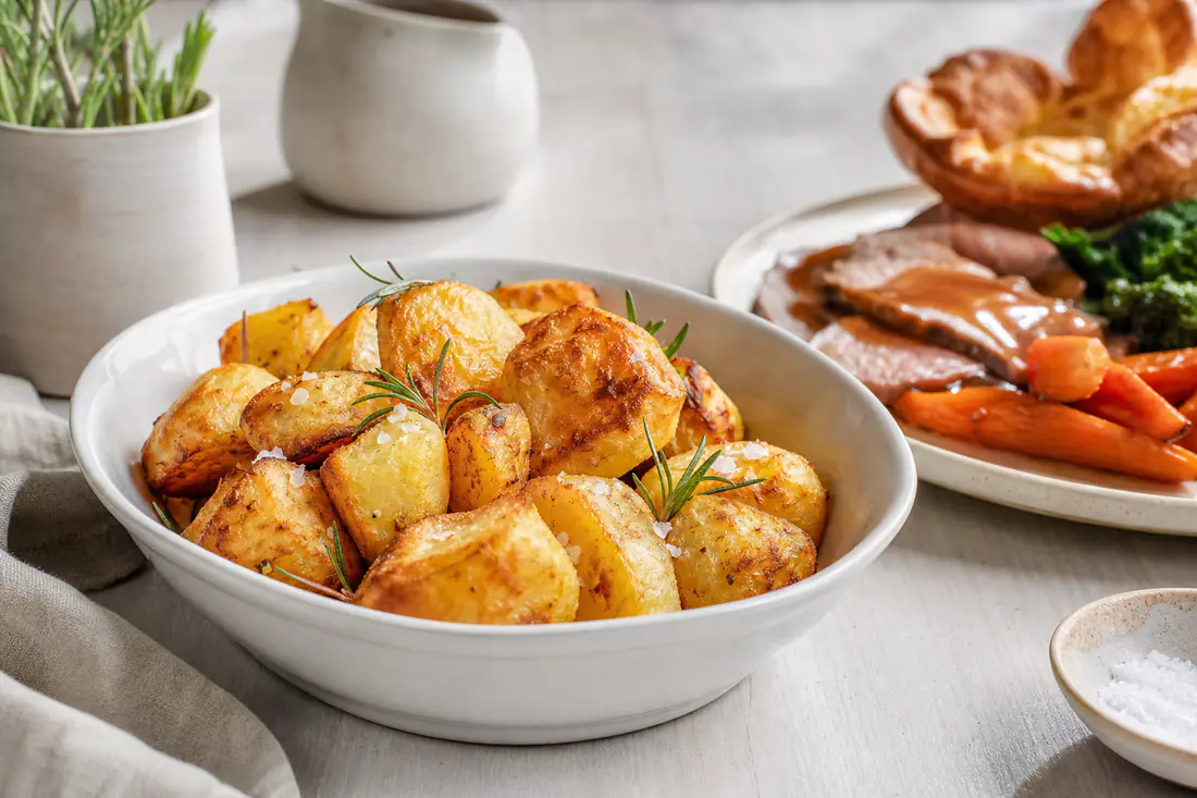 A white serving bowl of golden crunchy roast potatoes with rosemary beside a Sunday roast plate with gravy