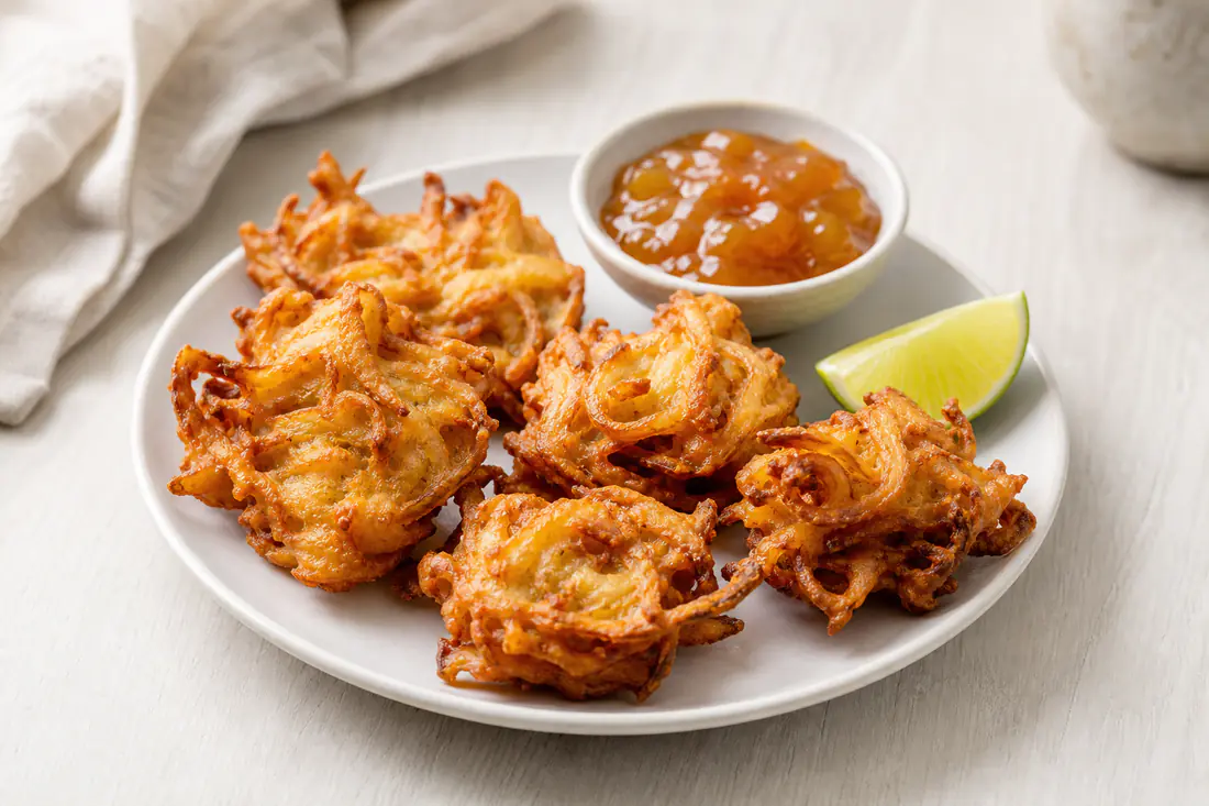 Four golden crisp onion bhajis on a plate with mango chutney and a wedge of lime