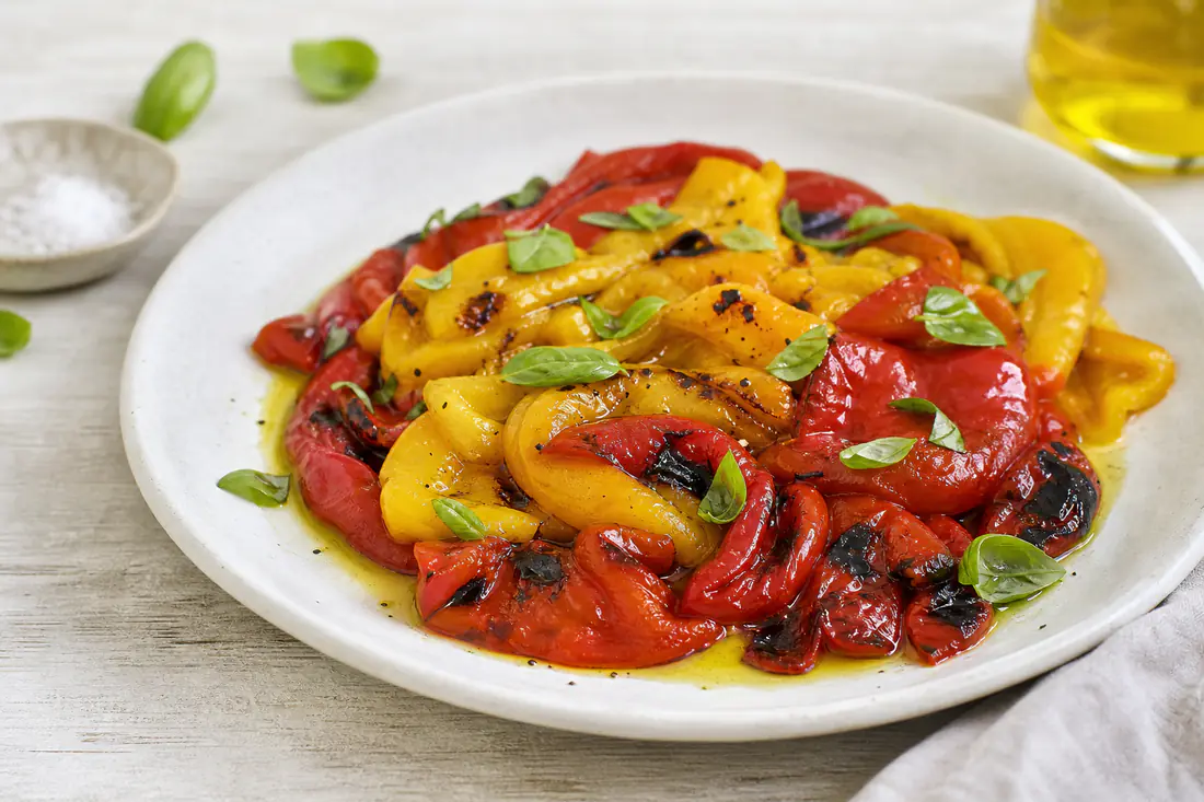 Charred red and yellow roasted peppers, some peeled and sliced into strips, on a white plate with olive oil and basil