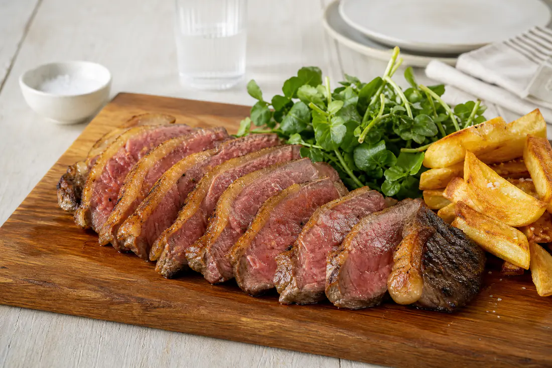 A sliced medium rare rump steak with a browned crust on a wooden board with chips and watercress