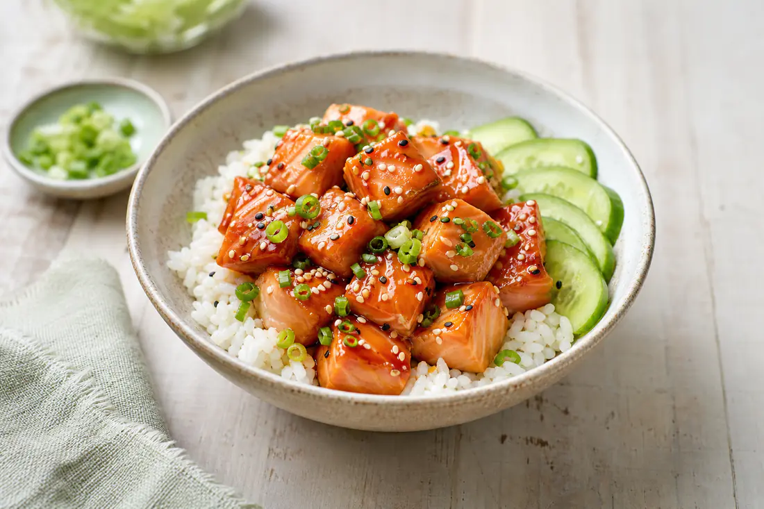 Glazed salmon bites with sesame seeds and sliced spring onion in a bowl of rice with cucumber
