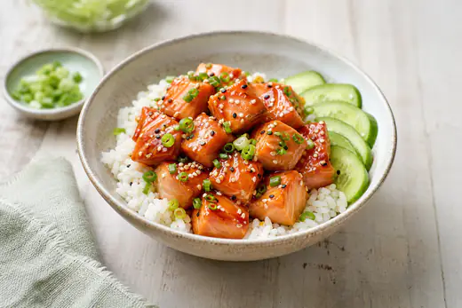 Glazed salmon bites with sesame seeds and sliced spring onion in a bowl of rice with cucumber