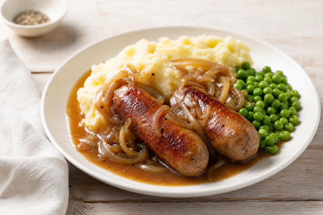 Browned pork sausages cooked from frozen on a plate with mashed potato, peas and onion gravy