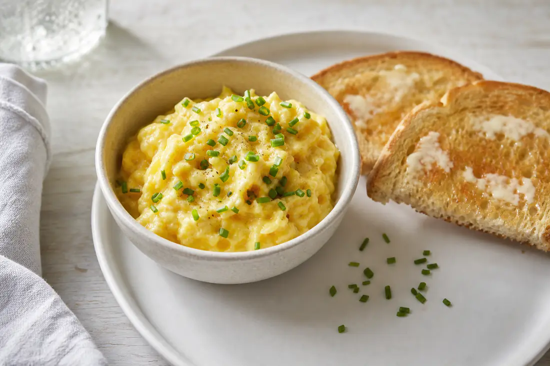 Soft scrambled eggs in a small ceramic dish next to buttered toast and chopped chives on a white plate