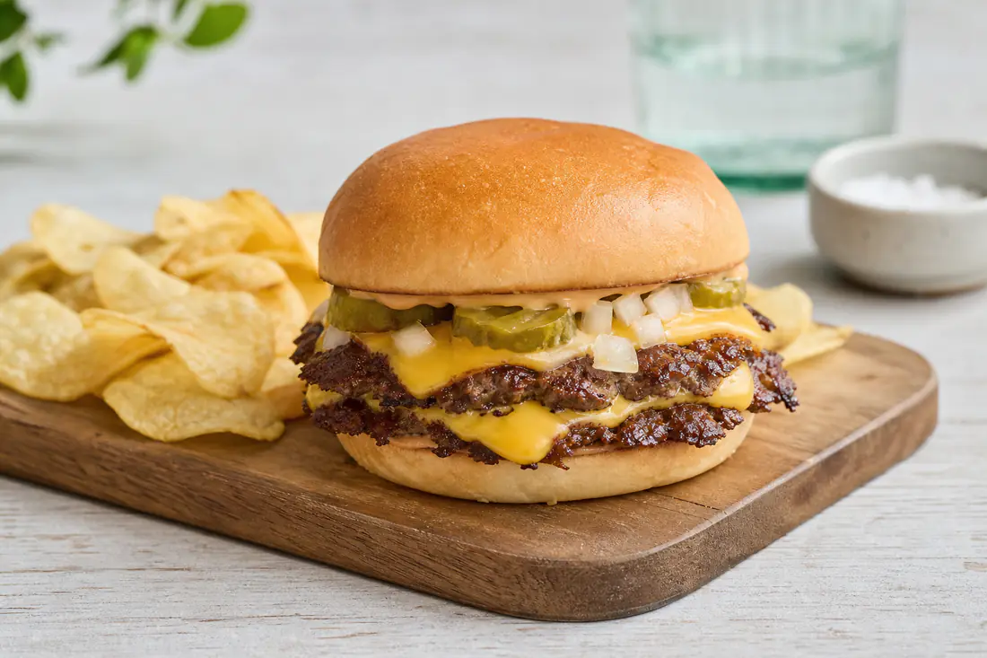 A double smash burger with melted cheese, pickles and onion in a soft bun on a wooden board