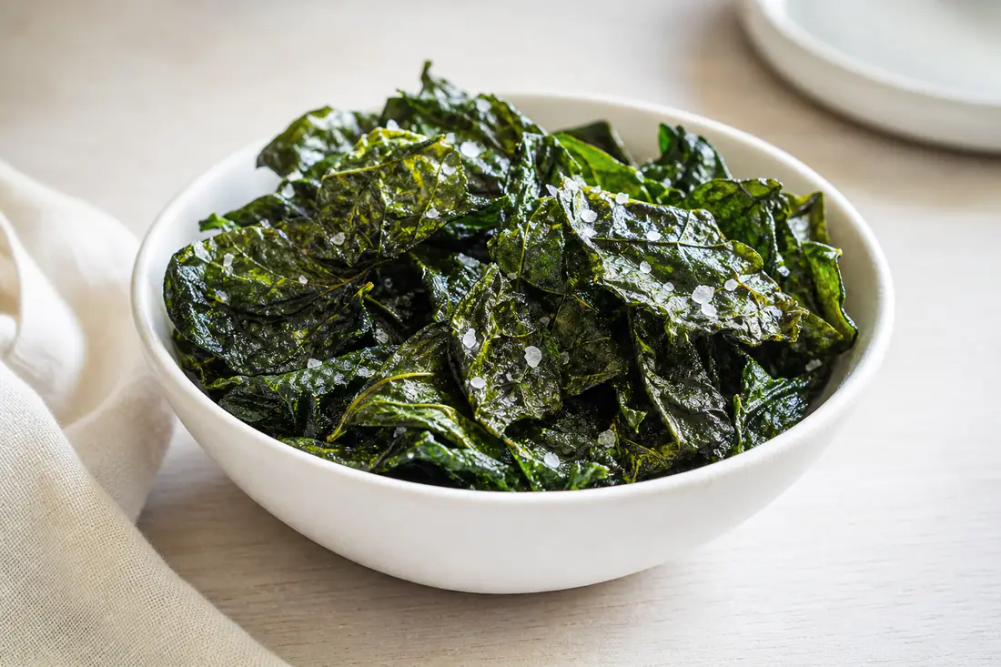 A white bowl of thin crispy dark green spinach chips lightly sprinkled with sea salt