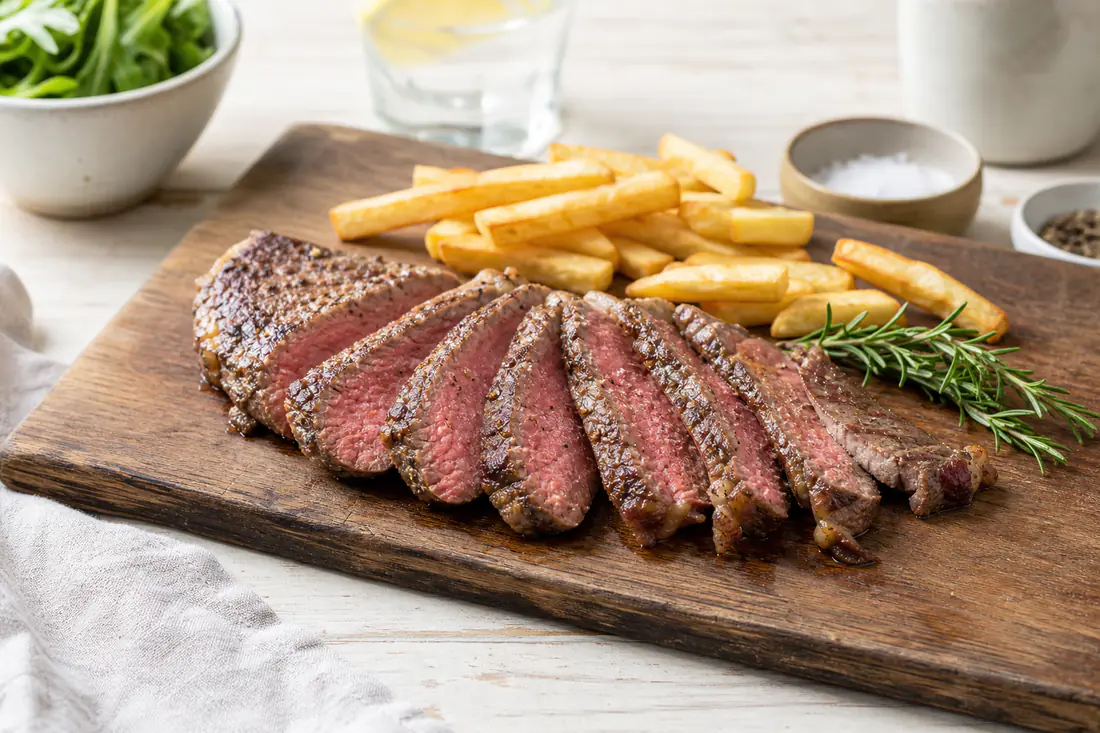 Sliced rump steak, browned outside and medium-rare inside, on a wooden board with chips and a sprig of rosemary
