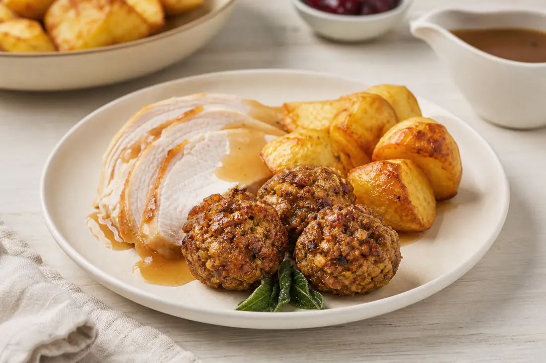 Golden brown sage and onion stuffing balls on a white plate beside roast turkey slices and gravy