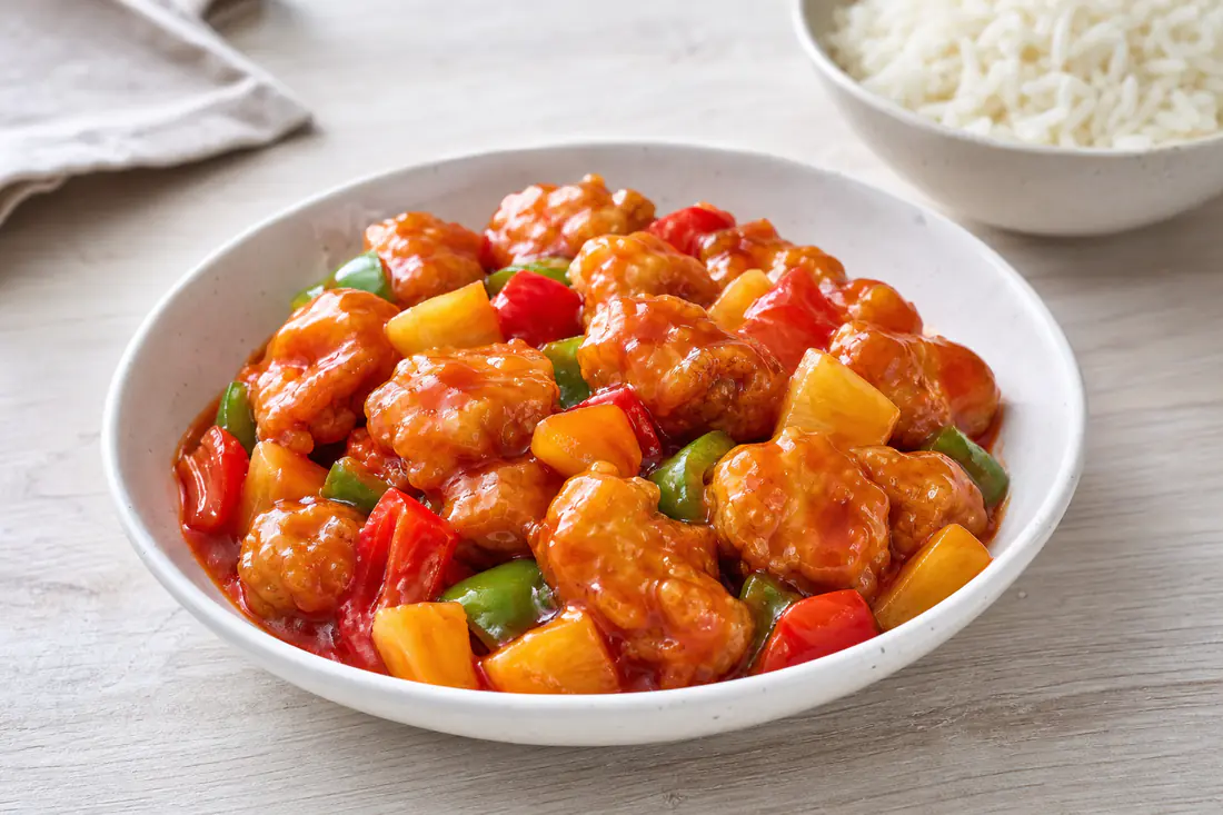 Crispy chicken pieces in glossy sweet and sour sauce with pineapple and peppers beside a bowl of rice