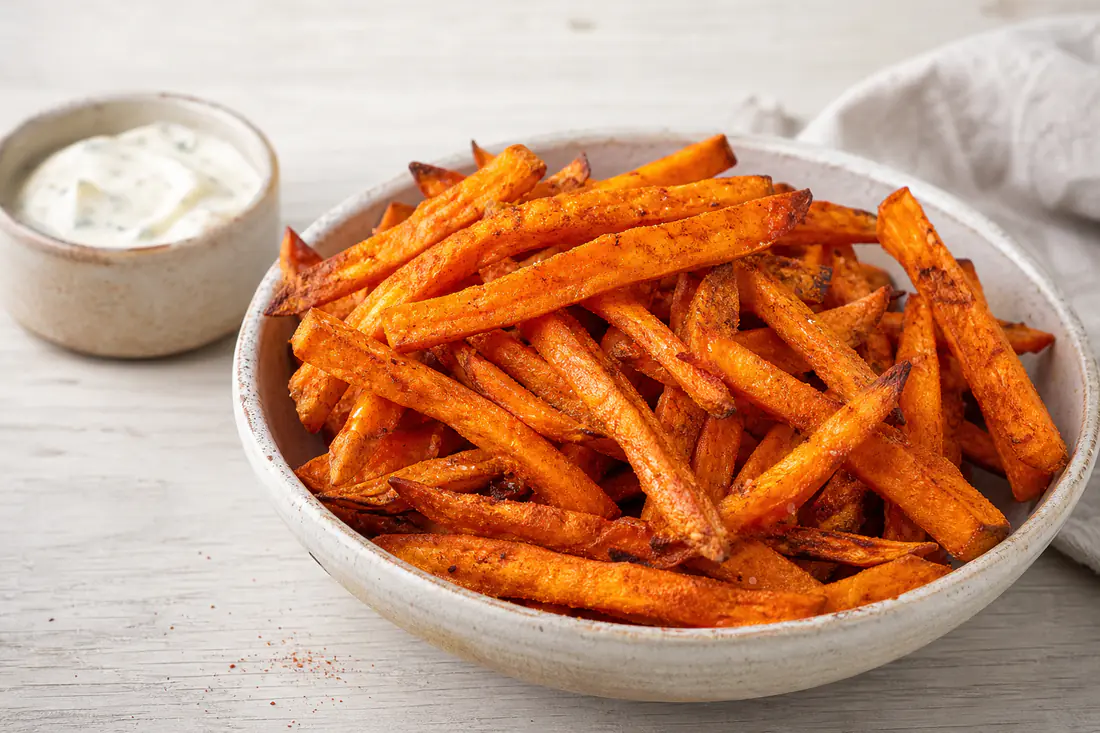 A pile of crisp orange sweet potato fries with smoked paprika in a bowl, with a garlic dip