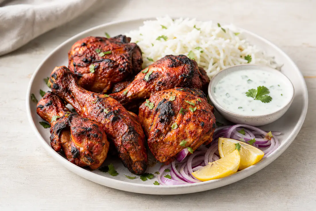 Red-orange tandoori chicken legs with charred edges on a plate with lemon wedges, onion rings and mint yoghurt
