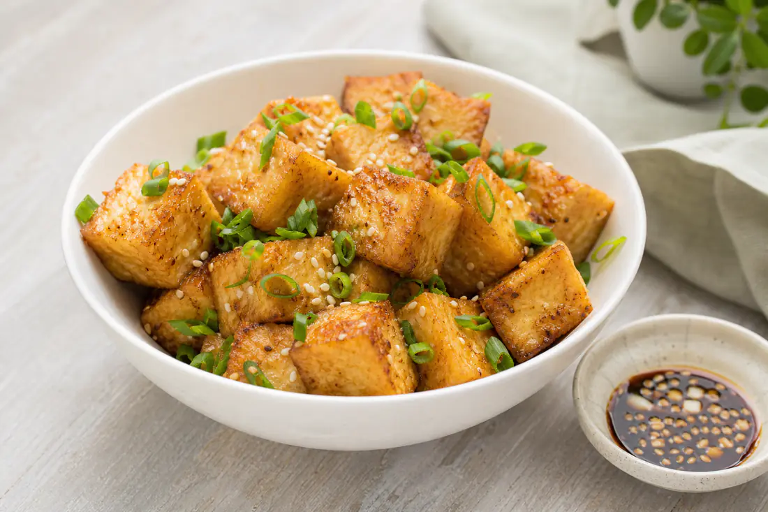 Golden crispy tofu cubes in a white bowl with sliced spring onions and sesame seeds beside a small dish of dipping sauce