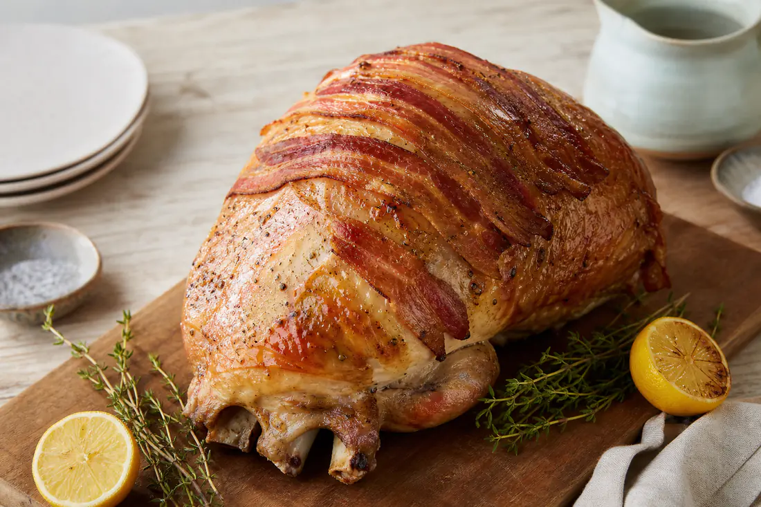 A golden roast turkey crown topped with crisp bacon rashers on a carving board with thyme and lemon halves