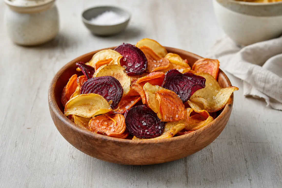 A wooden bowl of mixed homemade crisps made from potato, purple beetroot and orange parsnip slices with sea salt flakes